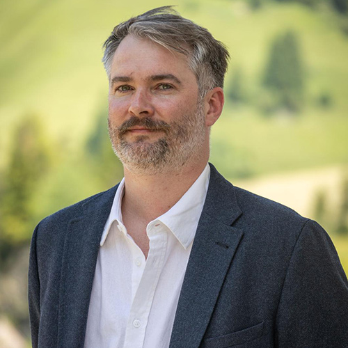 Headshot of a person with short grey hair and a beard wearing a blazer outdoors.