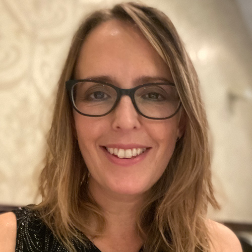 Headshot of a person with glasses and shoulder-length brown hair, against a blurred background.