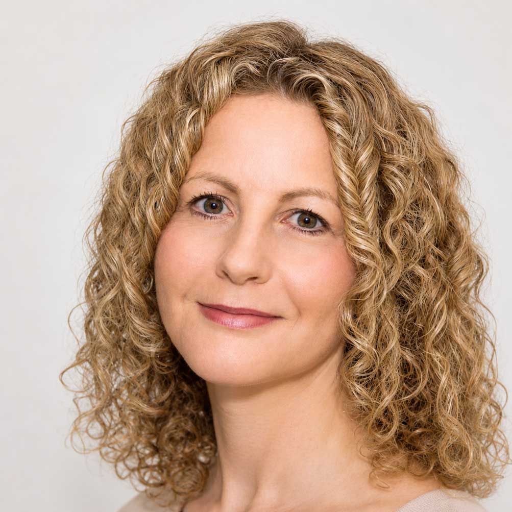Headshot of a person with shoulder-length, curly blonde hair smiling against a plain background.