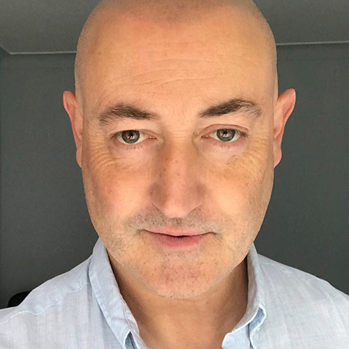 Headshot of a person with a shaved head, wearing a light blue shirt against a grey background.